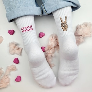 Könnte beinhalten: Ein Paar weiße gerippte Socken. Die linke Socke zeigt den gestickten rosa Text "ICH HASSE MENSCHEN" und die rechte Socke zeigt eine gestickte braune Hand, die ein Friedenszeichen macht. Die Socken werden zu einer hellblauen Jeans getragen.
