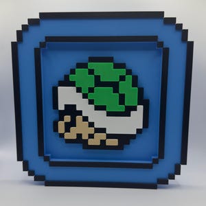 May include: A framed wall art piece with a pixelated design of a green and white turtle shell. The artwork is set within a blue frame with a black border, evoking a retro video game style.