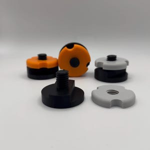 May include: A selection of black, orange, and grey camera accessories. The collection includes a threaded base, several circular components, and a few pieces with a notched design. The items are arranged against a plain white background.