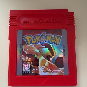 May include: A red Game Boy Color cartridge featuring a holographic image of a Pokemon character on the front. The cartridge is labelled "Pokemon" and has a "This Side Out" label on the right side. The ESRB rating is "Everyone" and the game is published by Nintendo.