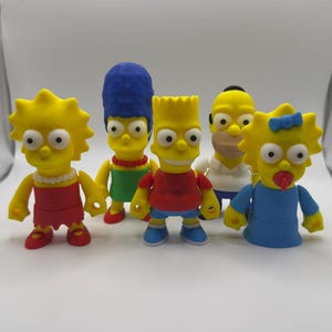 May include: Six cartoon character figurines with yellow skin and distinctive outfits. The figures are dressed in red, blue, green, and white. The characters have exaggerated features and are grouped together, possibly representing a family or team.