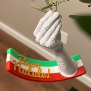 Puede incluir: Un adorno impreso en 3D de color blanco con forma de mano que sostiene una pancarta con la frase italiana "Buon Natale!" en letras doradas. La pancarta es roja, blanca y verde, asemejándose a la bandera italiana.