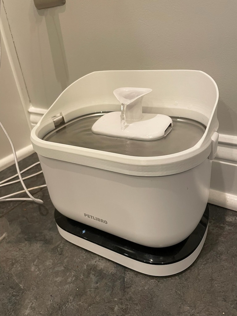 May include: A white PETLIBRO pet water fountain with a black base. The fountain has a water reservoir and a white spout with water flowing from it. Designed to provide fresh water for pets.