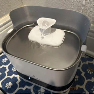 May include: A white and grey pet water fountain with a stainless steel basin. Water flows from a white spout into the basin. The fountain sits on a blue patterned mat.