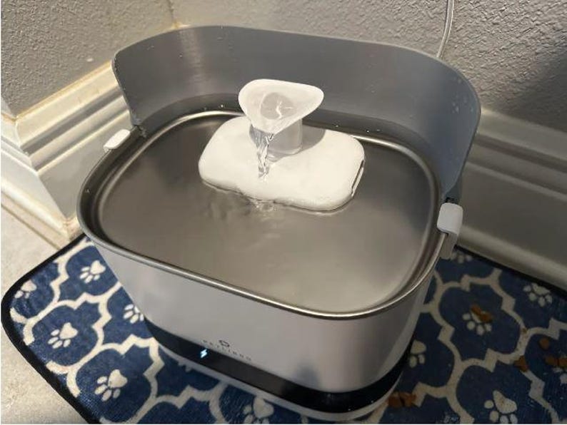 May include: A white and grey pet water fountain with a stainless steel basin. Water flows from a white spout into the basin. The fountain sits on a blue patterned mat.