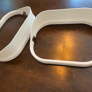 May include: Two white, 3D-printed, oval-shaped frames. One frame is a complete oval, while the other is a partial oval. The frames have a smooth, matte finish and are designed with a slight curve. The objects are placed on a dark wooden surface.