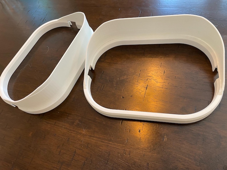May include: Two white, 3D-printed, oval-shaped frames. One frame is a complete oval, while the other is a partial oval. The frames have a smooth, matte finish and are designed with a slight curve. The objects are placed on a dark wooden surface.