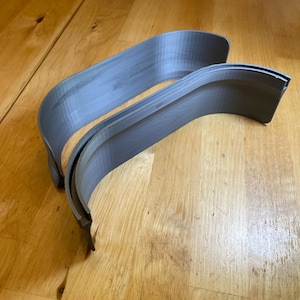 May include: Two grey, curved plastic components, possibly for a vehicle, rest on a wooden surface. The pieces have a layered texture and a smooth, rounded form. The lighting accentuates the curves and the material's subtle variations.