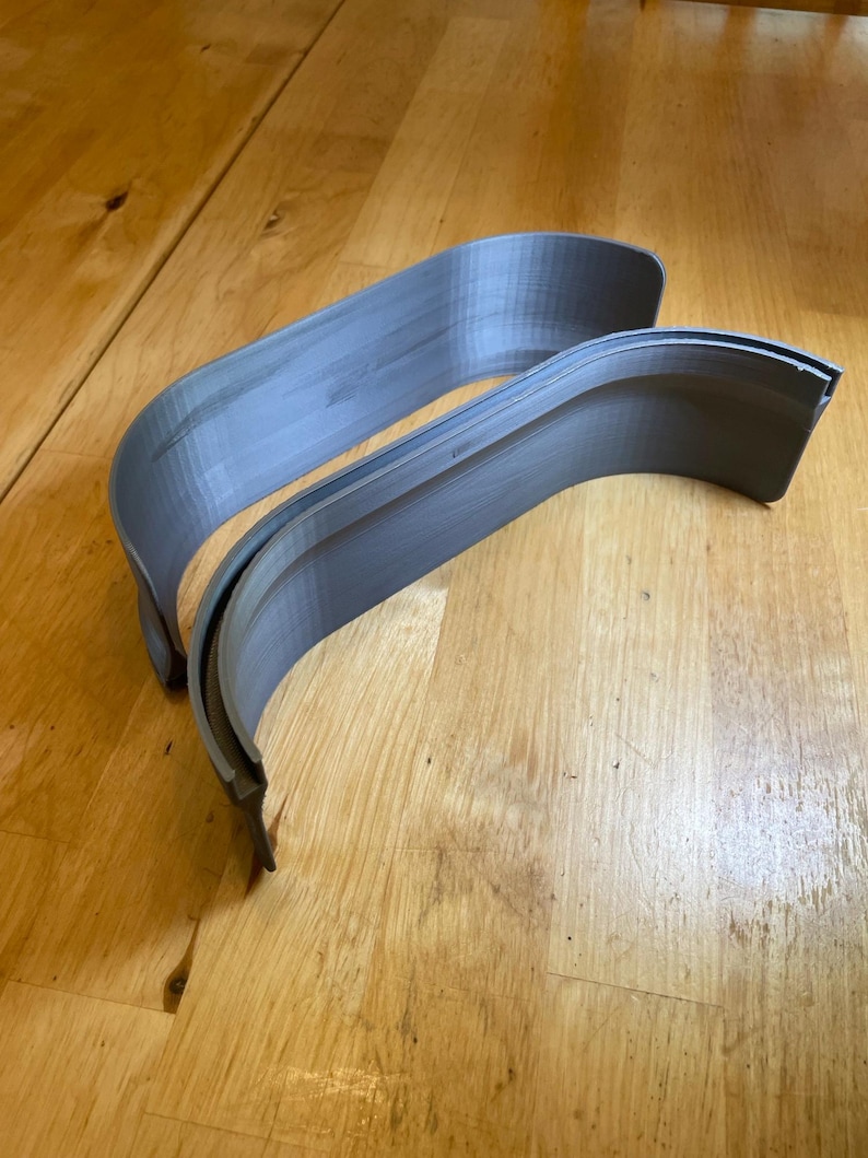 May include: Two grey, curved plastic components, possibly for a vehicle, rest on a wooden surface. The pieces have a layered texture and a smooth, rounded form. The lighting accentuates the curves and the material's subtle variations.