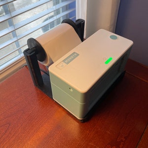 May include: A white and light blue label printer with a roll of white label paper. The printer has a green light on the front and the word "MUNBYN" printed on the side.