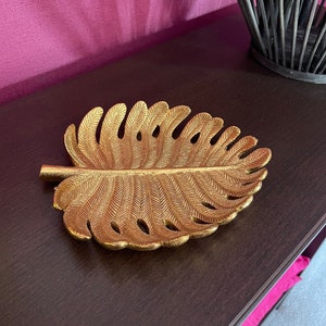 May include: A gold leaf-shaped decorative tray with a textured surface. The tray is on a dark brown surface.