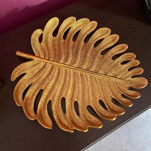 May include: A gold leaf-shaped decorative tray with a detailed design. The tray is made of metal and has a shiny finish.