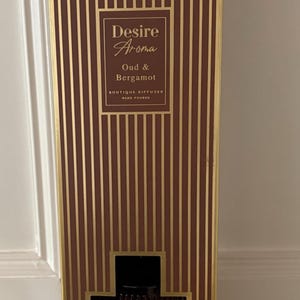 May include: A brown and gold striped rectangular box with the text "Desire Aroma Oud & Bergamot". The box is a boutique diffuser, with a dark brown base and gold trim.