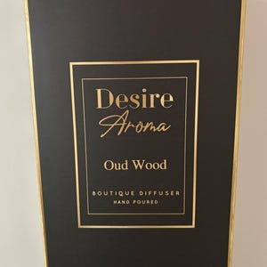 May include: Black rectangular box with gold trim and a gold framed label. The label reads "Desire Aroma Oud Wood" with "Boutique Diffuser Hand Poured" below. The box is for a home fragrance product.