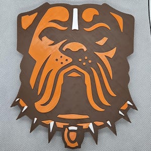 May include: A flat, wall-mounted decorative plaque shaped like a bulldog head. The design features a dark brown base with layered orange accents forming the facial features and a spiked collar. Small white details highlight the forehead and collar spikes.