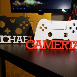 May include: Black and white gaming controller-shaped desk decorations. One features the name "MICHAEL" in white letters, and the other has the word "GAMERTAG" in red letters. The controllers are on a dark surface.