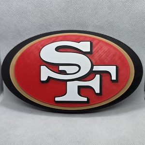 May include: An oval-shaped wall plaque featuring the San Francisco 49ers logo. The design consists of a white interlocking SF monogram centered on a red oval background, bordered by a thin gold ring and a wider black outer oval frame.