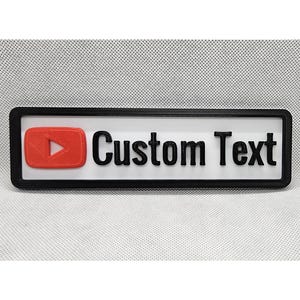 May include: A rectangular sign with a black frame, white background, and the words "Custom Text" in black. A red play button icon is on the left. The sign is likely 3D-printed.