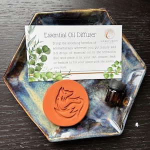 May include: An essential oil diffuser set includes a hexagonal ceramic dish with blue and brown glaze, a terracotta disc with a fox design, and a small brown bottle. A card reads "Essential Oil Diffuser".