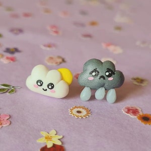May include: A pair of handmade polymer clay earrings featuring a cute smiling white cloud with a yellow sun and a sad grey cloud with raindrops.