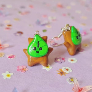 May include: A pair of dangle earrings featuring a brown clay character with a green leaf on its head. The leaf has a smiling face with black eyes and a black line down the center.