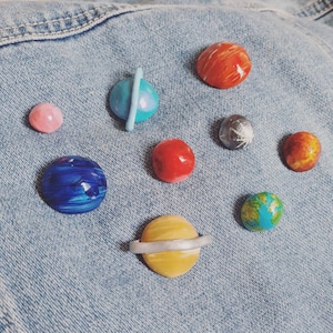 May include: A set of nine planets made of polymer clay, each painted with different colors and details. The planets are arranged on a blue denim background.