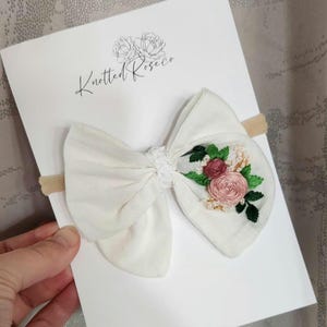 May include: A white fabric bow headband featuring an embroidered floral design with pink roses and green leaves on one side. The bow is attached to a tan elastic band and displayed on a white card with the text Knotted Roseco and a floral logo.