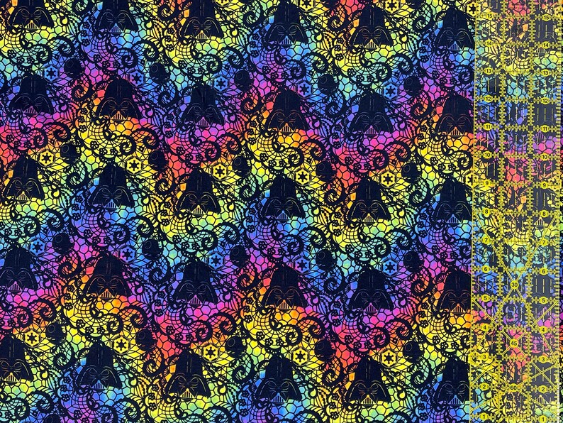 May include: A patterned fabric featuring black Darth Vader helmets and floral lace against a rainbow gradient. A clear ruler with yellow markings is on the right. Ideal for crafting and sewing. The fabric's design is eye-catching and suitable for various projects.