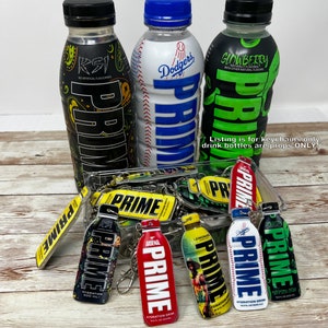 Puede incluir: Una colección de llaveros de botellas de bebida Prime Hydration de colores vivos. Los llaveros presentan diferentes sabores y diseños, incluyendo KSI, Dodgers, Glowberry, Lemonade, Arsenal e Hydration Green. Los llaveros se muestran sobre una superficie de madera con botellas de bebida vacías en el fondo. La oferta es solo para los llaveros. Las botellas de bebida son solo accesorios.