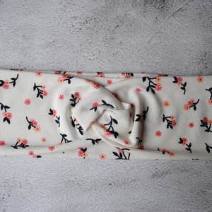 Könnte beinhalten: Cremefarbenes Stirnband mit floralem Muster. Der Stoff ist mit kleinen rosa Blüten und schwarzen Blättern bedeckt. Das Stirnband ist in der Mitte verdreht und schafft ein stilvolles Accessoire.