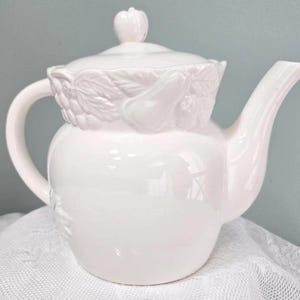 May include: A white ceramic teapot with a lid and handle. The teapot features a decorative relief of leaves and fruit around the lid. The spout is curved, and the handle is a simple loop. The teapot is sitting on a white lace tablecloth.