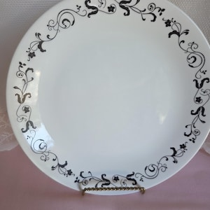 May include: White ceramic plate with a black floral design around the rim. The plate is round and has a glossy finish. The floral pattern includes stylized leaves and flowers.