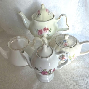 May include: Four ceramic teapots in various shapes and sizes. The teapots are primarily white with floral designs in pink, blue, and other colors. Some have gold trim. The teapots are arranged on a white lace surface.