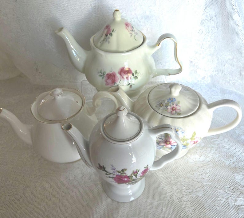 May include: Four ceramic teapots in various shapes and sizes. The teapots are primarily white with floral designs in pink, blue, and other colors. Some have gold trim. The teapots are arranged on a white lace surface.