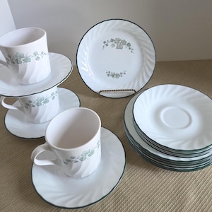 May include: White ceramic teacups and saucers with a green floral design and a green trim. The set includes several cups, saucers, and plates. The plates have a spiral design.