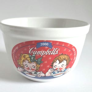 May include: White ceramic Campbell's soup bowl with a red and white label featuring two children eating soup. The label includes the Campbell's logo and the year 2000. A vintage collectible.