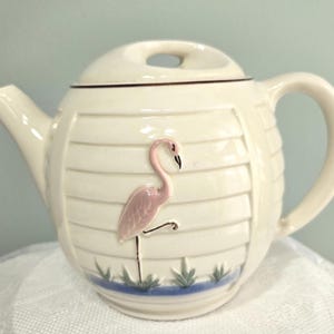 May include: Cream-colored ceramic teapot with a lid and handle. The teapot features a pink flamingo design on the front, set against a background of horizontal lines. The base has a blue and green design. The teapot is designed for brewing tea.