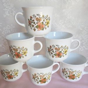 May include: A stack of six white ceramic mugs with a floral design in shades of orange, yellow, and brown. Each mug has a rounded shape with a handle. The mugs are arranged on a pink surface.