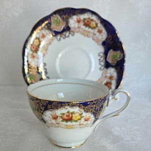 May include: A white teacup and saucer set with a floral and gold design. The cup and saucer have a dark blue border with gold accents. The cup has a handle and a gold rim. The saucer has a scalloped edge.