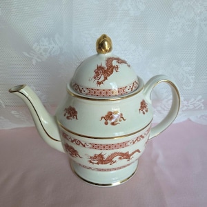 May include: Cream-colored teapot with a lid, featuring a gold finial and gold trim. The teapot is adorned with red dragon and cloud motifs. The handle and spout are elegantly curved, offering a classic design.
