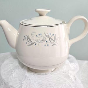 May include: A white teapot with a silver trim and a floral design in shades of gray and blue. The teapot has a round body and a lid with a knob.
