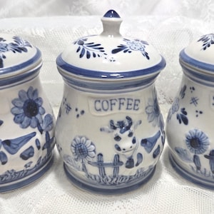 May include: Three ceramic canisters with lids, labeled "TEA", "COFFEE", and "SUGAR". Each white canister has blue floral and cow designs. The lids and rims are also blue. The canisters are likely for kitchen storage.