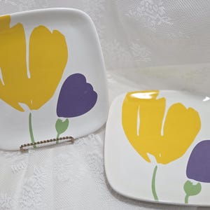 May include: Two square white ceramic plates with a yellow tulip design and a purple accent flower. The plates have a green stem and are set against a white background.