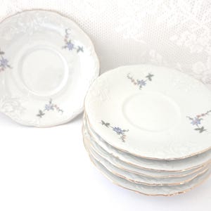 May include: A stack of six white porcelain saucers with a scalloped edge and gold trim. Each saucer features a delicate floral design in blue, pink, and green. One saucer is separate, and the background is white.