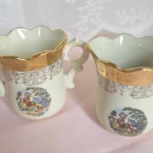 May include: Two antique cream-colored porcelain cups with gold trim and ornate handles. Each cup features a decorative scene with figures in a garden setting. The rims have a scalloped design.