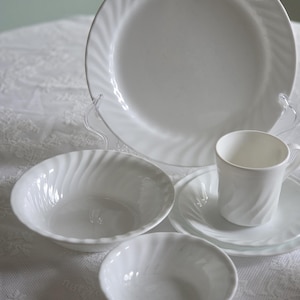 May include: A white ceramic dinnerware set featuring a large plate, two bowls, a saucer, and a teacup. Each piece has a spiral design around the rim. The set is displayed on a white lace tablecloth.