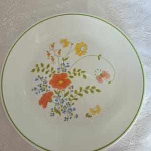 May include: White ceramic plate with a green rim and a floral design. The design features orange, yellow, and blue flowers with green leaves. The plate is round and has a simple, elegant aesthetic.