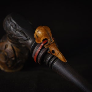 May include: A wooden pipe with a carved bird head design. The bird head is made of a light brown wood and has a red and black leather strap around it.