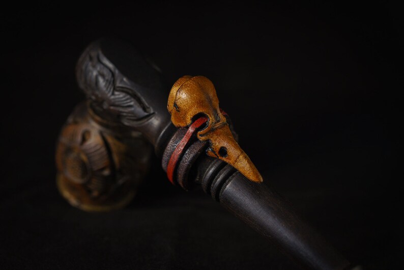 May include: A wooden pipe with a carved bird head design. The bird head is made of a light brown wood and has a red and black leather strap around it.