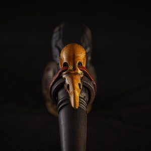 May include: A wooden pipe with a carved bird head design. The bird head is painted in shades of brown, yellow, and red. The pipe is decorated with a leather strap.